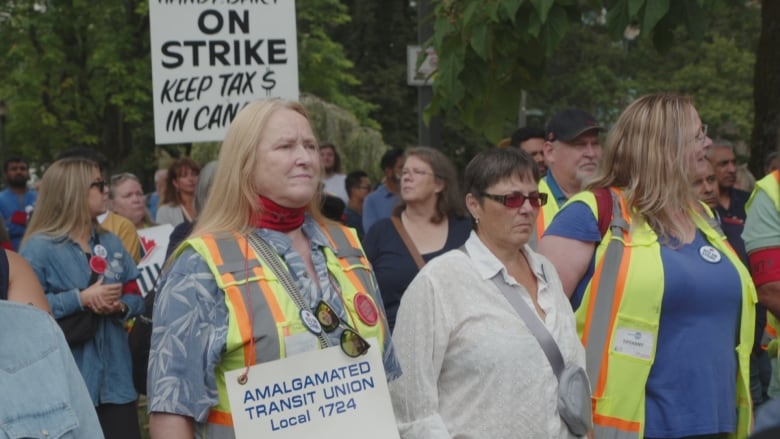 A number of people are seen at a rally. One of them holds a sign that reads 'On Strike Keep Tax $ In Can'.