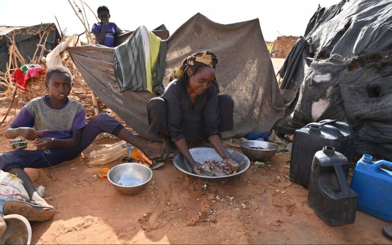 A woman sits on the ground washing clothes in a metal bowl surrounded by a shelter wall made from twigs and cloth. A young boy sits beside her. Another boy is seen standing in the background. 