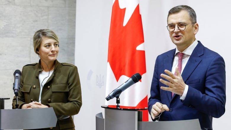 A man and a woman dressed formally are shown at individual platforms at what appears to be a news conference. The bespectacled man is speaking and gesturing as the woman looks on.