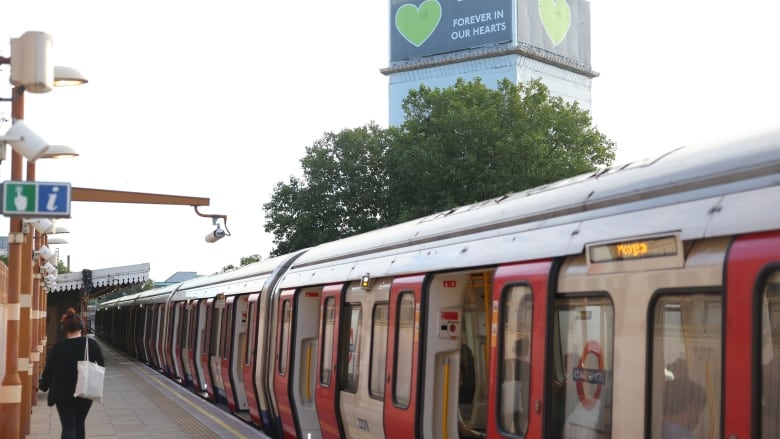 In the foreground, people on a subway platform are shown as a train sits on the tracks. In the distance, a high-rise residential building is shown.