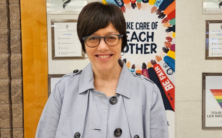 A woman with dark hair and eyeglasses stands in front of the door to a school classroom.