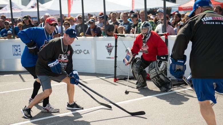 In daylight, at a street hockey rink with boards, three players fight over an orange ball in front of a goalie and net. Fans are packed against the boards, watching the action.