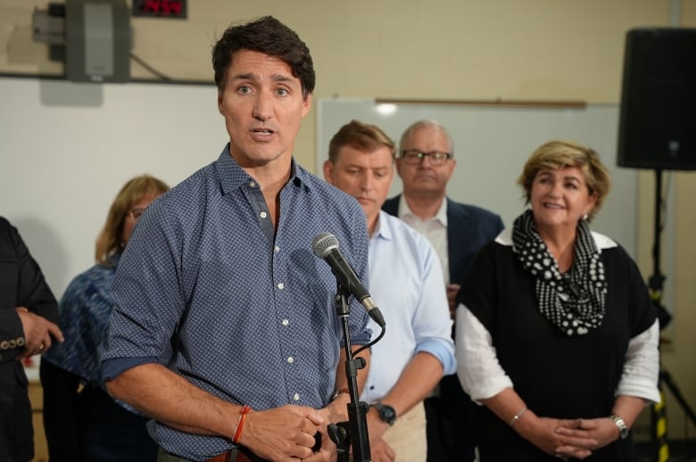 A group of people stand in a classroom. One man stands in front of them speaking at a microphone