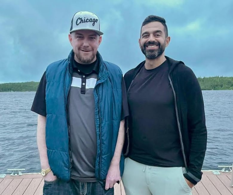 Two smiling men stand next to each other on a pier, water behind them.