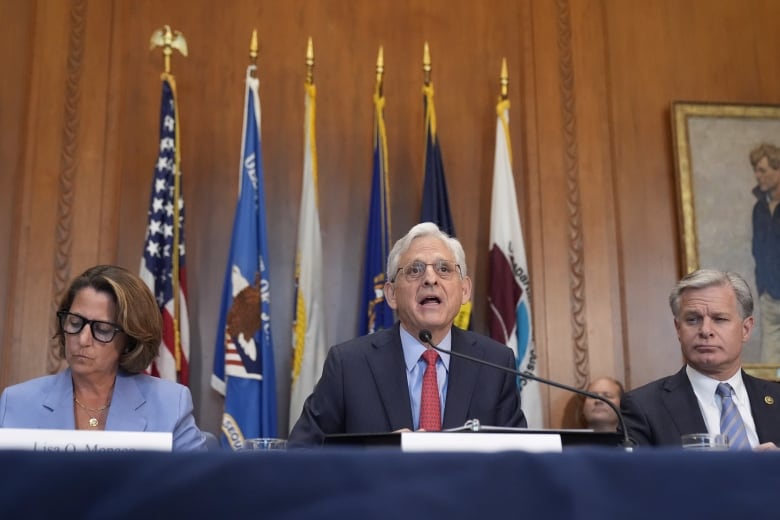 A woman in a lavender suit, a man in a grey suit and a man in a charcoal suit sit in a row at a table covered in a navy blue tablecloth. Black microphones are in front of each person.