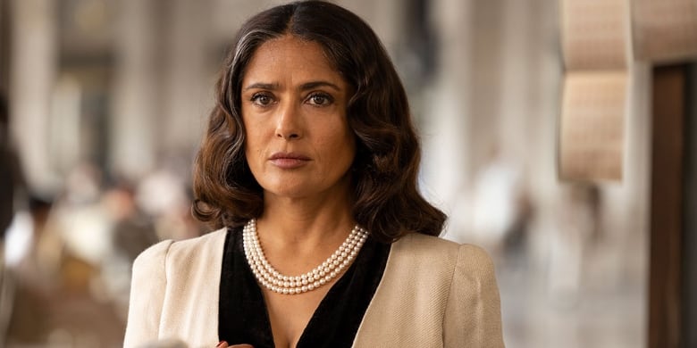 Salma Hayek, a Mexican-American woman stands wearing a white blazer and white pearls.