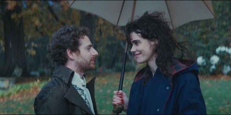 A white man and woman face one another as the woman holds an umbrella, surrounded by trees. 