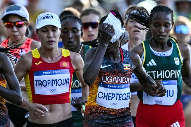 A woman marathon runner, with a bib on her chest bearing the name Cheptegei, holds a bag of ice against her face as she runs among a crowd of other women.  