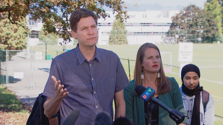 David Eby, a tall figure with dark hair, wearing a short-sleeved patterned shirt gestures in front of microphones with a playing field behind him. A woman with blonde hair and wearing bright red lipstick stands beside him. A girl stands beside them.