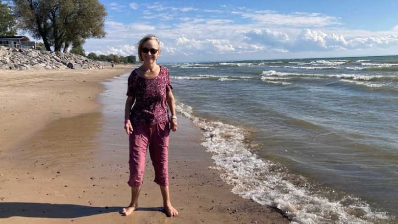 A woman wearing sunglasses, standing on a sandy beach as waves roll in.