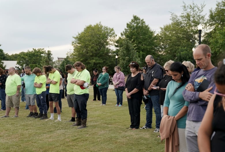 Over a dozen people are shown in casual wear, some with heads bowed, on a field.