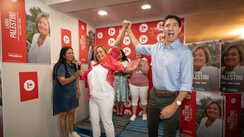 A man in a blue button up shirt raises the hand of a woman wearing a white shirt and red overshirt. People in the background applaud.