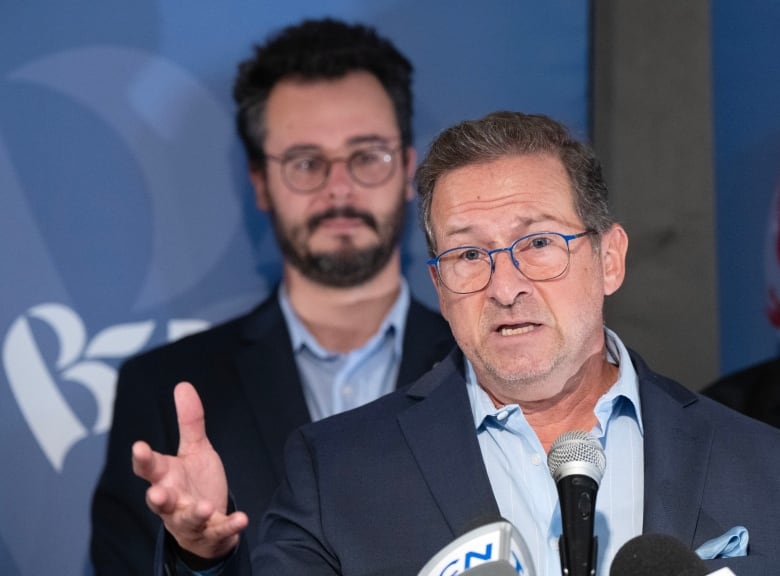 A man in glasses and a suit speaks into a microphone as another man looks on.