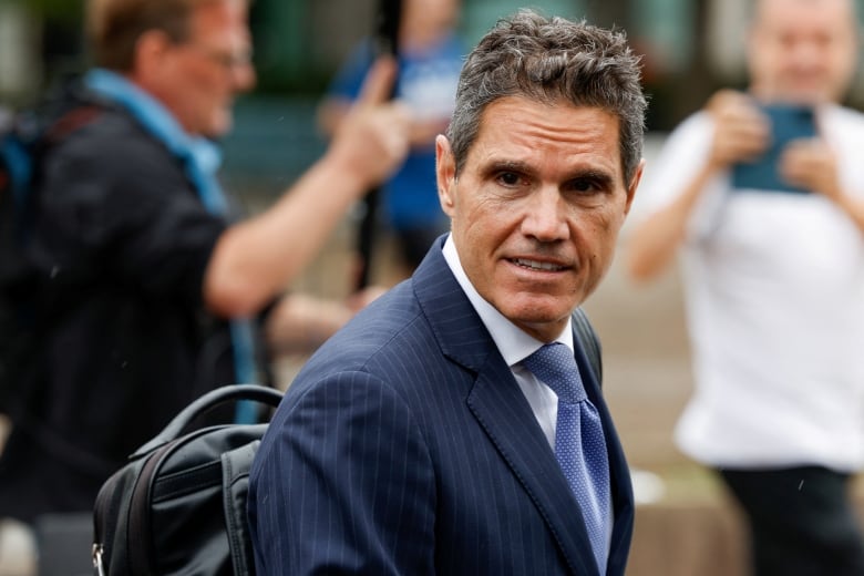 A dark-haired clean shaven man in a suit and tie is shown in closeup.