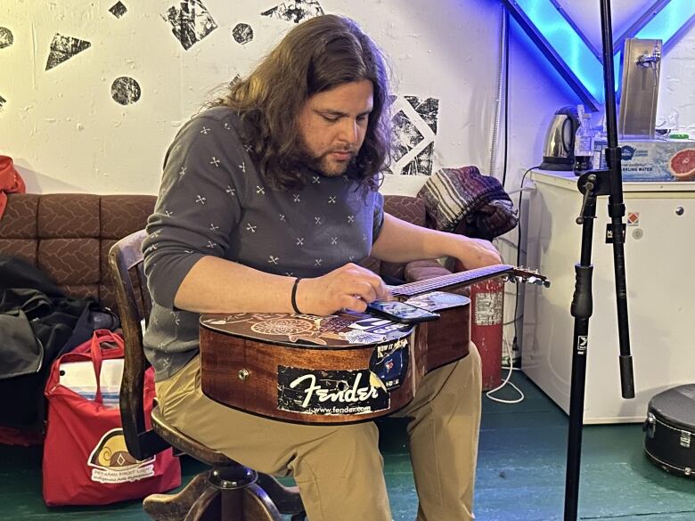 A person with shoulder-length, wavy brown hair sits with a guitar on their lap.