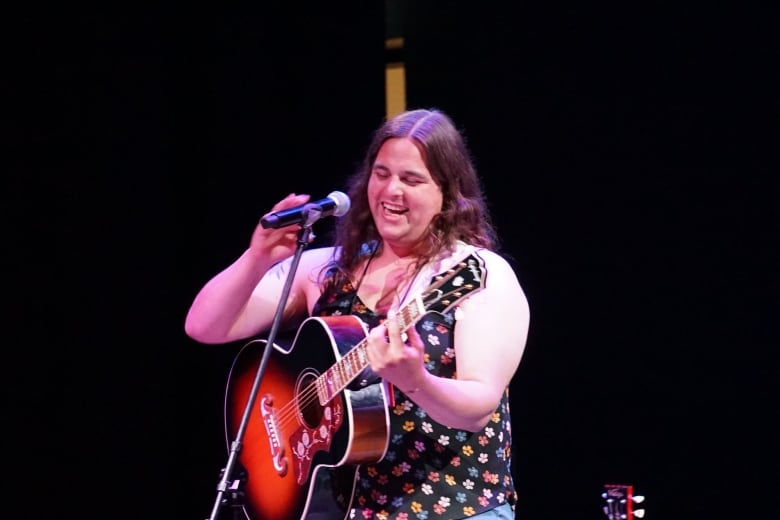 A woman with long brown hair and wearing a flower-patterned dress stands smiling in front of a microphone, holding a guitar.