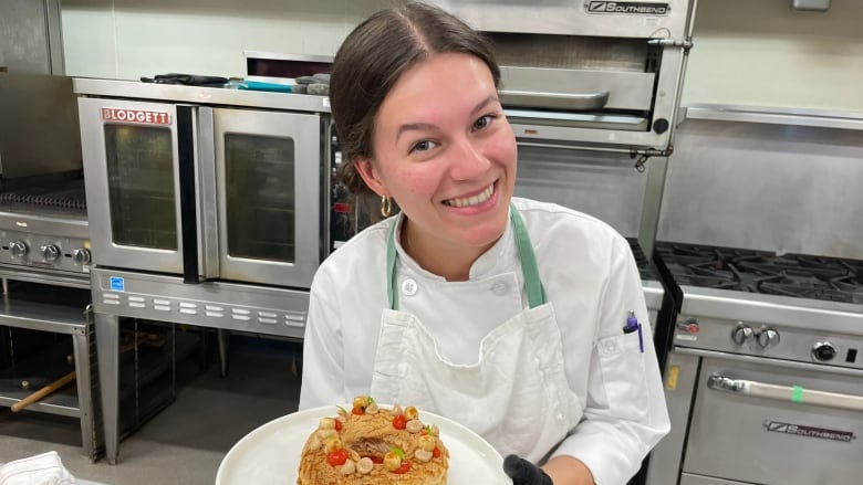 A woman wearing chef attire holds a plate with a small cake on it. 