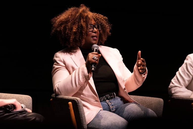 A woman in jeans and a white blazer speaks into a microphone while sitting in a chair. 