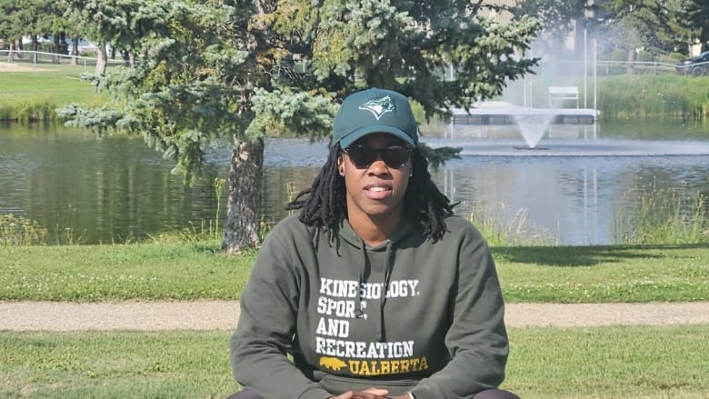 A Black woman sits on a park bench in the sunshine, behind her is a green field and a small lake with a fountain.