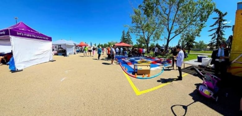 A number of tents are set up on a paved pad on a sunny blue day. There are games set up and crowds walking around.