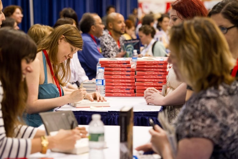 A woman in a crowded room sits next to a stack of hardcover novels signing the cover pages. 