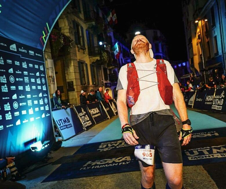 A man is wearing a red vest, shorts and a headlamp as he crosses the finish line of a race. he is looking up to the sky. He has red hair and a beard.