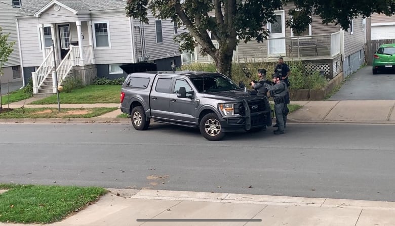 Three officers dressed in swat gear and holding long guns stand next to a grey pickup truck.