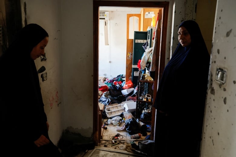 Two women wearing head coverings stand near the door of a heavily damaged room of what appears to be a residential dwelling, with debris and items strewn about the ground.