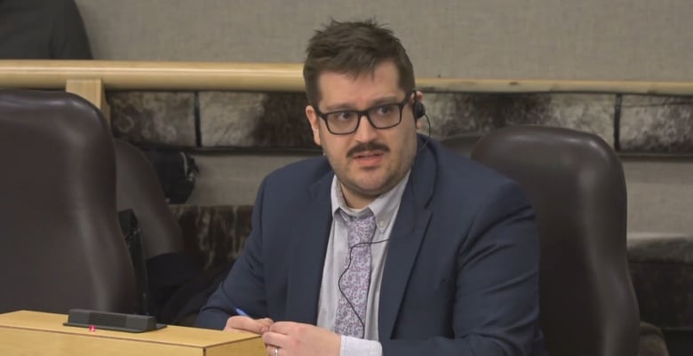 A man wearing a blue shuit, grey shirt, purple tie and glasses sitting in a legislative room wearing an earpiece