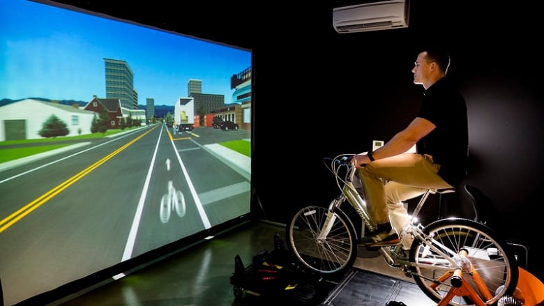 A man on a bicycle in a dark room with a screen displaying a simulated streetscape in front of him.