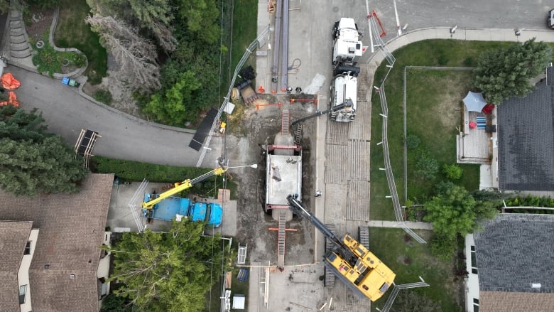 Large construction machines are parked near an excavated section of road, exposing a large pipe.