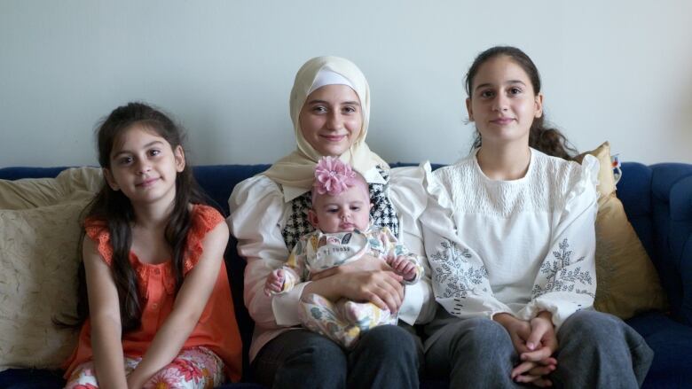 Three girls of differing ages sitting on a couch while smiling at the camera, the girl in the middle is holding a baby sitting on her legs.