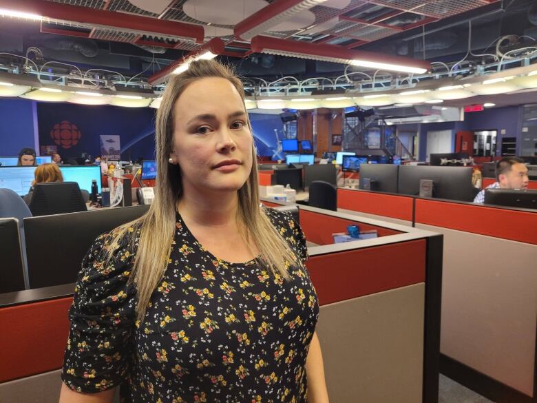 A blond woman stands in a black blouse in a newsroom. 