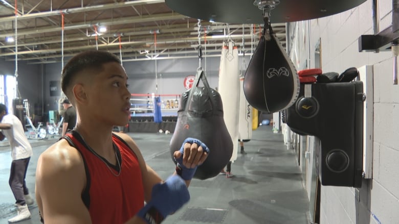 A boy wearing a red sleeveless shirt with his hands taped takes a fighting stance in front of a black speedbag.