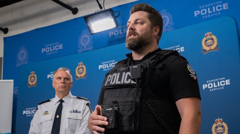 A uniformed police officer is pushing a button on a device on his vest. It lights up red. He is standing in front of another officer in a conference room.