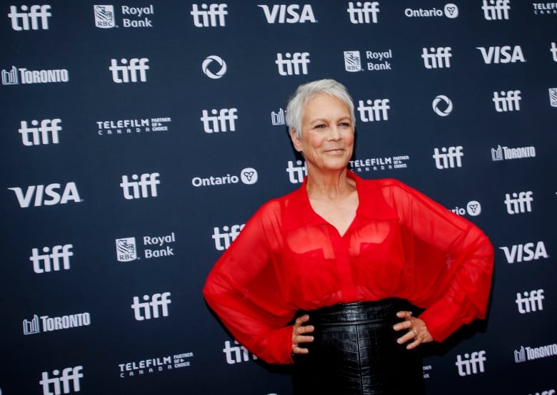 A woman with grey hair, wearing a red blouse and a black skirt, poses for a photo in front of a dark blue backdrop with the words 