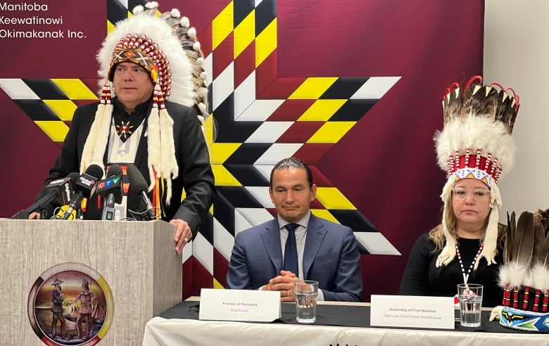 A man in an Indigenous headdress speaks at a podium, with a man and woman sitting at a table next to him.