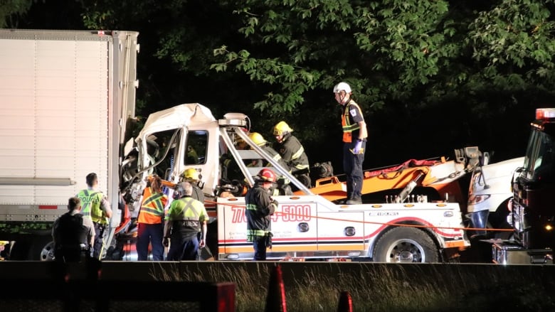 First responders wearing high-vis gear gather around a destroyed tow truck on a road.