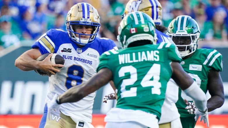 A quarterback in a blue and white uniform carrying a football faces two people on green and white uniforms.