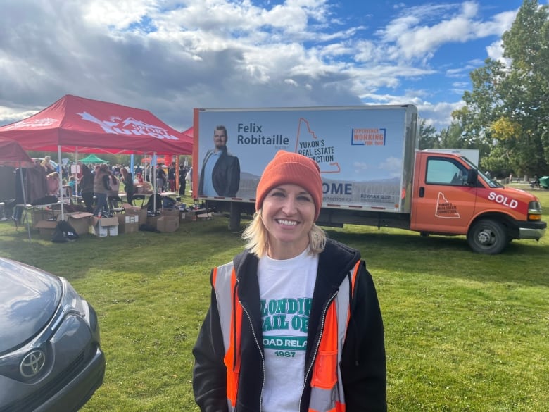 A woman in an orange toque and an orange vest with blonde hair stand in front of an orange moving truck.