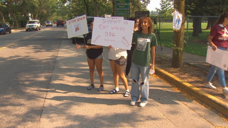 A number of young people in the street hold signs advertising a BBQ and car wash.