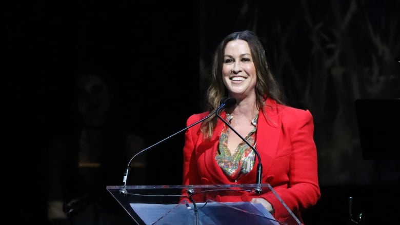 Alanis wears a red suit while speaking at a clear podium.