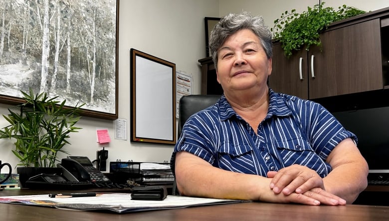 A woman with light skin and short grey hair is wearing a blue shirt with white vertical stripes. She is sitting, with her hands folded, at an office desk.