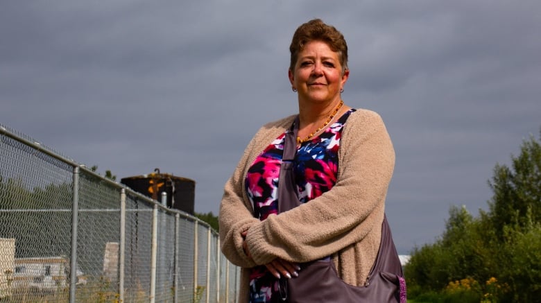 A woman with short auburn hair is wearing a colouful top, under a beige cardigan. A maroon leather bag drapes across her shoulder. She is standing outside, her hands together, beside a chain-link fence.