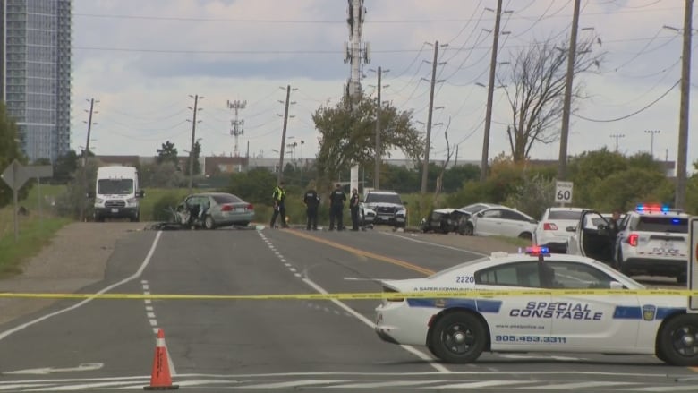 A road is taped off and police cars are on scene next to two smashed up sedans on a grey day. The road is surrounded by telephone poles and greenery