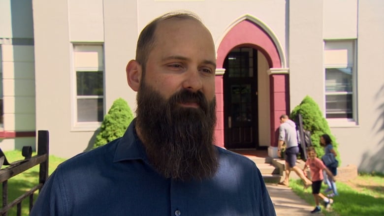 Man with bald head and long brown beard wears navy collared shirt in front of a school.