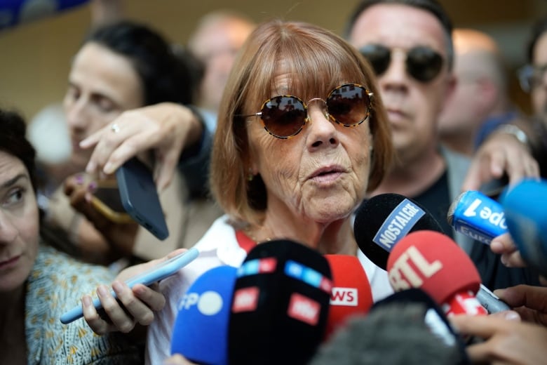 An  older woman speaks into several press microphones in a scrum