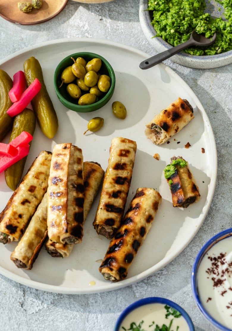 Overhead shot of a platter of grilled pitas stuffed with kebabs. Green olives and assorted pickles sit next to them. 