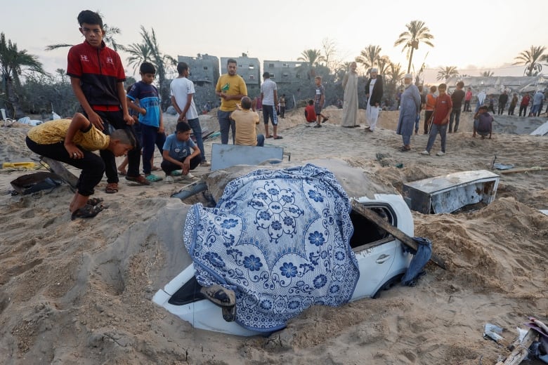 Children peer into a white car that is buried in the sand.