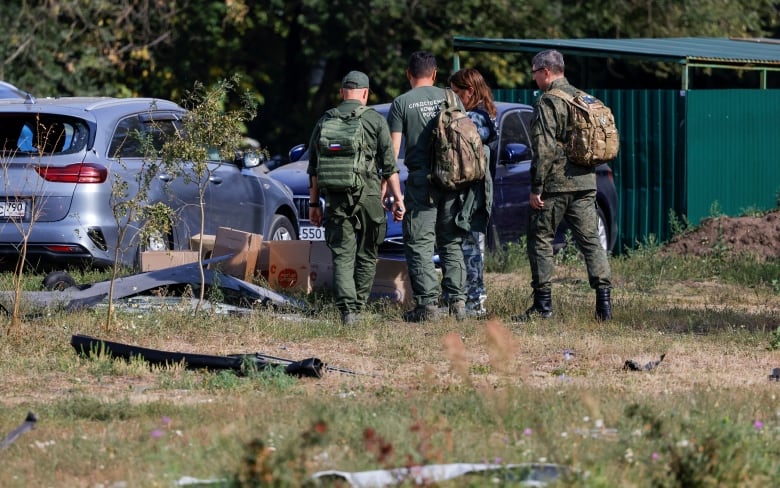 Several officials in uniform are shown in a grassy area near a damaged vehicle. 